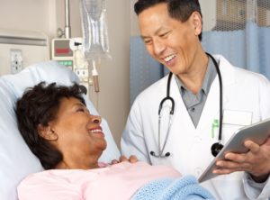 A picture of a doctor wearing a white lab coat and a stethoscope around his neck smiling at a patient who is laying in a hospital bed.