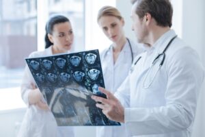 Three Doctors in White Lab Coats Wearing Stethoscopes Around Their Neck Talk About a Photo of a Brain Scan One Doctor is Holding