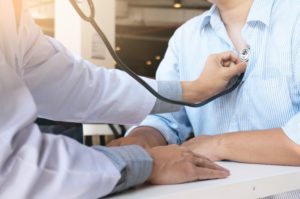 A picture of a male doctor wearing a stethoscope and holding it to a patient’s chest. 