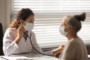 A picture of a doctor wearing a mask, a white lab coat, and a stethoscope, listening to a patient’s heart who is also wearing a mask.