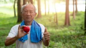 Elderly man wearing a white shirt with a blue towel around his shoulders, holding a heart and giving a thumbs up while staying fit with a walk in the woods.