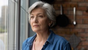 Older woman with grey hair wearing a blue denim shirt and staring out of a window.