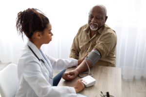 A Doctor in a White Lab Coat Wearing a Stethoscope Around Her Neck Takes an Older Male Patient’s Blood Pressure at a Wood Table