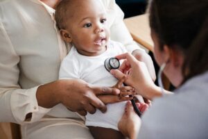 A baby sitting on the lap of a woman in all white smiles and looks at a physician who has a stethoscope to his heart 