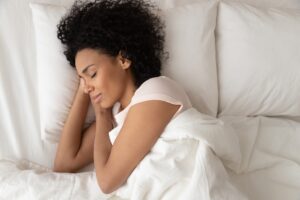 A Woman Smiles While Sleeping Peacefully on Her Side Under the Covers in White Bedding with Her Hands Under Her Head