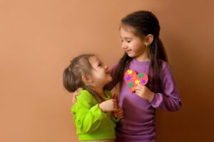 A little girl with her arm around a smaller girl, smiling at each other and holding paper hearts. 