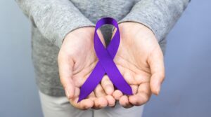 The Torso of Someone in White Pants and a Grey Sweatshirt with Upright Hands Held Out and a Purple Ribbon Resting in Them