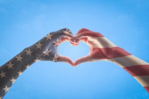 Arms that look like the American Flag coming together to form the shape of a heart in front of a blue sky.